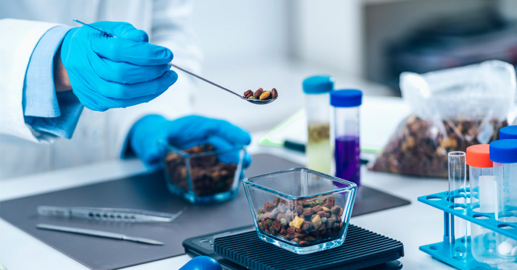 Gloved hands in a lab putting pet food into a glass container on a scale.