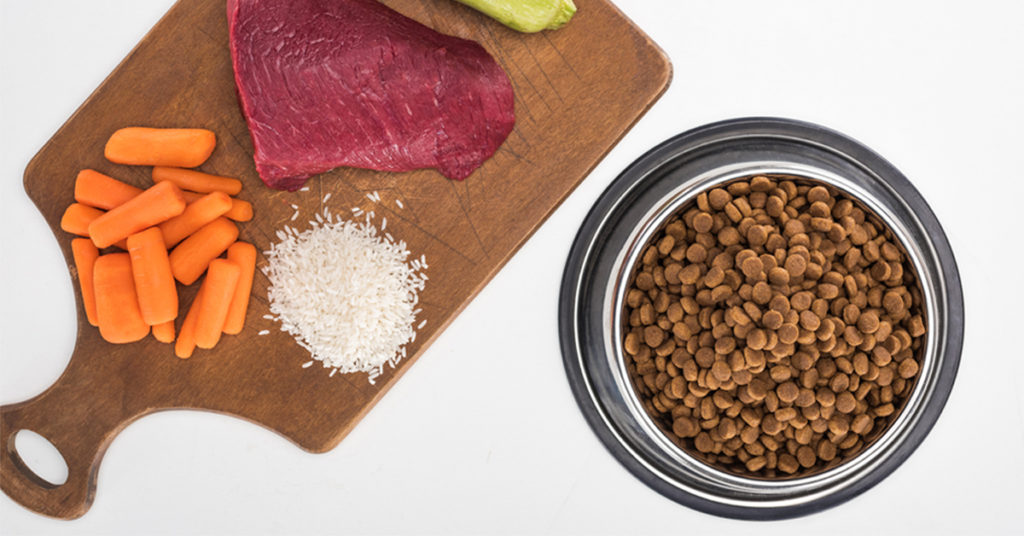 A wood cutting board with carrots, beef, and rice next to a bowl of pet food.