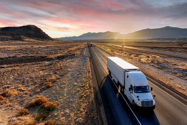 Semi truck driving down a road with the sun setting behind mountains.